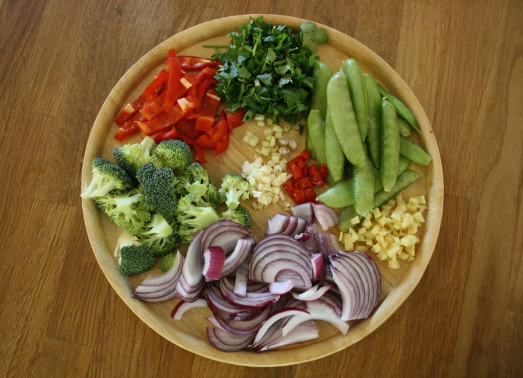 wooden plate with cut vegetables  in piles including peppers, onion, broccoli and sugar snap peas.