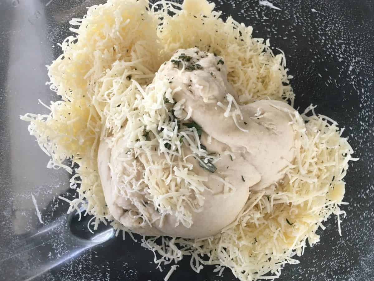dough in a glass bowl with grated cheese and herbs on top.