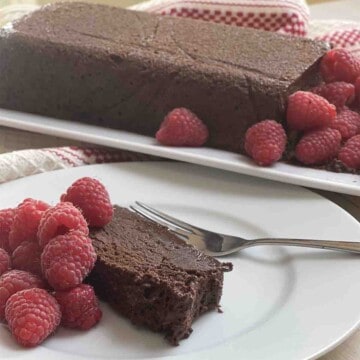 slice of chocolate marquise on a plate with raspberries and the rest of the dessert in the background.